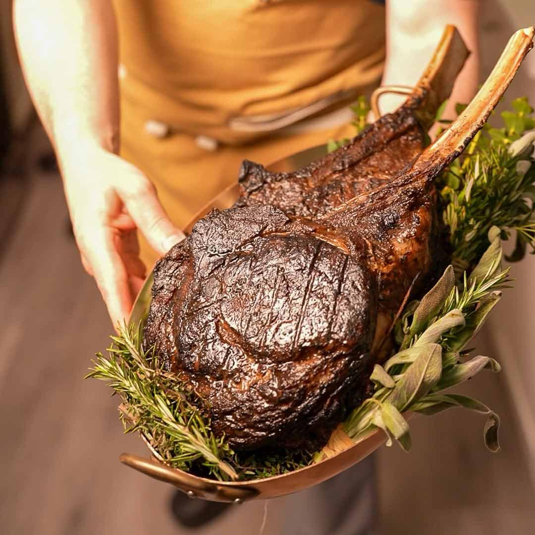 Grilled tomahawk steak served on a platter with fresh herbs, held by a server.